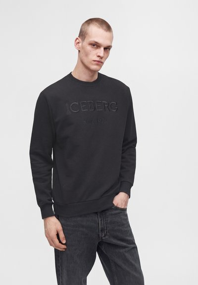 Jeune homme portant un sweat-shirt noir avec le texte "ICEBERG since 1974" et un jean gris foncé, debout devant un fond uni clair.