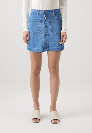 Lys blå denimnederdel med en tætsiddende talje, frontknaplukning, to store påsyede lommer og en rullet kant. Bæres med beige slidesandaler.