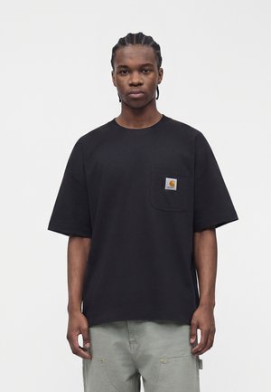 Jeune homme avec des tresses cornrows porte un t-shirt Carhartt noir avec une poche poitrine et un pantalon gris clair, debout devant un fond uni.
