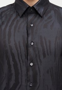 Μαύρο σατέν πουκάμισο με κουμπιά, με διακριτικό γυαλιστερό μοτίβο ζέβρας και μυτερό γιακά ορατό στον κορμό.