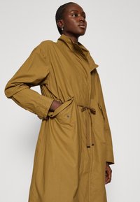 Trench coat lungo di colore giallo senape con colletto alto, cintura e tasche laterali, realizzato in un tessuto liscio e leggero.