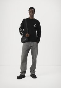 Pulover negru cu desene grafice pe mâneci, asortat cu blugi gri largi și încălțăminte chunky negre. Geantă de umăr purtată pe torso.