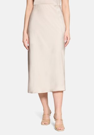 Kobieta ubrana w beżową satynową spódnicę midi i beżowe buty na obcasie z odkrytymi palcami, stojąca z luźno opuszczonymi ramionami na tle jednolitego tła.