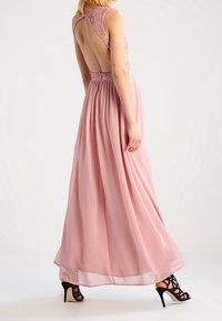 Maxikjol i mjuk rosa chiffong, med en djup ryggutskärning, dekorerad halsringning och en flödande kjol, tillsammans med svarta remsandaler.