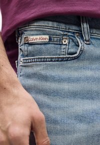 Άτομο που φοράει ανοιχτό γαλάζιο τζιν με ετικέτα Calvin Klein στην μπροστινή τσέπη και μπλου ζάκι μπορντό, το χέρι χαλαρό στο πλάι.