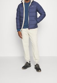 Marinblå puffjacka med en krämfärgad fleecefoder, tillsammans med en blå sweatshirt och elfenbenfärgade sweatshirtbyxor; svarta sportskor fulländar outfiten.