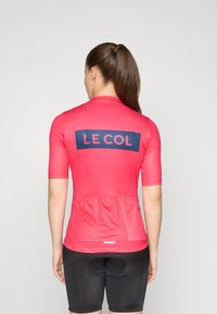 Kortermet sykkeltrøye i lys rosa med svart "LE COL" logo og et mesh baklommer. Brukt med svarte polstrede sykkelshorts.