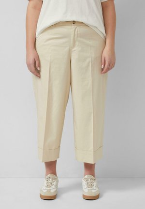 Persoon met beige cropped wijde broek en witte sneakers, staand tegen een effen lichtgrijze achtergrond.