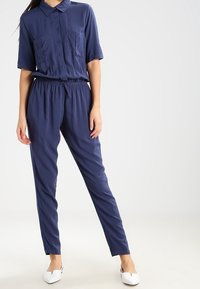 Marineblå jumpsuit i slät tyg, med krage, korta ärmar, två bröstfickor och resår i midjan.