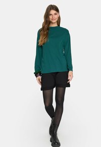 Zielona bluzka z długim rękawem w zestawieniu z czarnymi szortami. Modelka ma na sobie czarne rajstopy i botki za kostkę. Gładki materiał, luźny krój, zrolowane mankiety szortów.