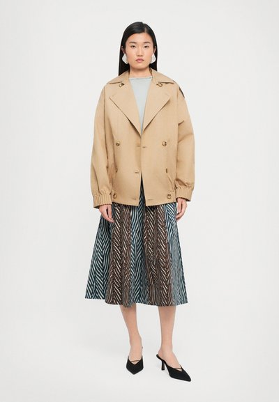Beige oversized dubbelknäppt jacka med stora gyllene knappar, bärs över en ljusgrå topp och en lång mönstrad kjol i blått, brunt och vitt.