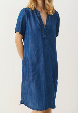 Femme portant une robe en denim bleu longueur genoux avec manches courtes, col en V, poches latérales et ourlet arrondi sur fond uni.