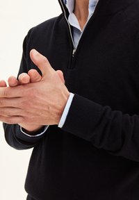 Sweat à zip noir avec des poignets côtelés. En dessous, une chemise rayée bleu clair et blanche est visible au niveau du col. La position des mains indique un geste de serrage.