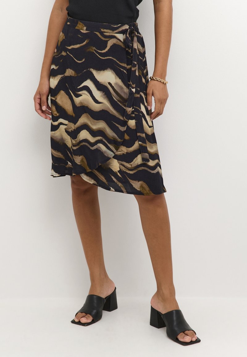 Femme portant une jupe portefeuille mi-longue à imprimé animalier foncé avec un lien sur le côté, un haut noir sans manches, des chaussures à talons carrés ouvertes à l'avant noires, et un bracelet en or.