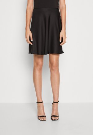 Femme portant une jupe noire jusqu'aux genoux, un haut noir sans manches, et des sandales à talons hauts à bride à la cheville, debout devant un fond uni.