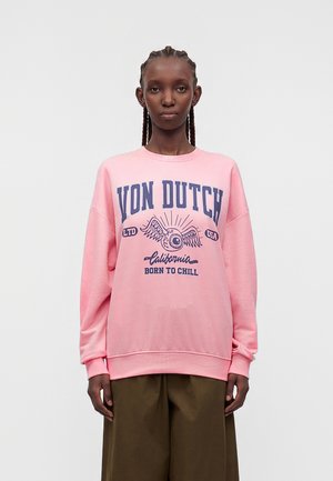 Persoon met gevlochten haar die een roze sweatshirt draagt met "VON DUTCH" en een vleugelachtig oog grafiek, gecombineerd met bruine wijd uitlopende broeken.