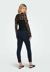 Top lungo nero in pizzo a maniche lunghe con motivo floreale, abbinato a jeans skinny scuri. Il top ha una silhouette aderente e un dettaglio trasparente sulla schiena.