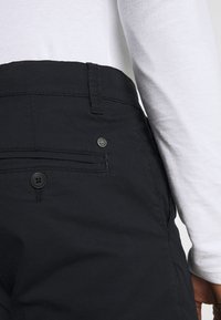 Pantalones negros de tela suave con un bolsillo trasero que tiene un cierre de botón y un pequeño detalle de logo. Parte de un atuendo informal con una camiseta blanca.