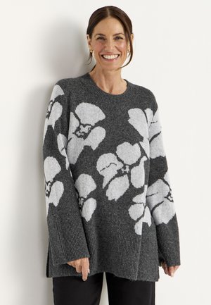 Vrouw glimlachend, draagt een donkergrijze trui met grote witte bloemenpatronen en een zwarte broek, staand tegen een effen lichte achtergrond.