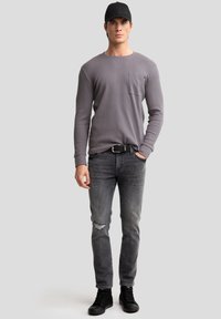 Camisa gris de manga larga con un bolsillo, combinada con jeans oscuros desgastados y zapatillas negras, completada con una gorra negra.