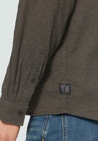 Grau gewebter Hemdenstoff mit einem strukturierten Muster; verfügt über Knopfmanschetten und ein kleines schwarzes Etikett mit weißem Logo am Saum.