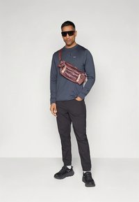 Camisola escura de manga comprida, calças pretas, ténis pretos. Bolsa de cintura Patagonia castanha com logótipo. Óculos de sol grandes. Modelo está em pé contra um fundo simples.