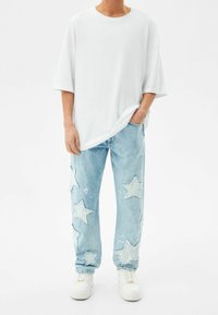 Persona vestida con una camiseta blanca de gran tamaño, jeans de mezclilla azul claro con grandes parches de estrella y zapatillas blancas, de pie contra un fondo blanco liso.