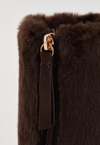 Barna műszőrmével készült táska, arany cipzárral és bőrkivitelű húzóval. A textúrája puha és bolyhos, hangsúlyozva gömbölyded formáját és meleg színét.