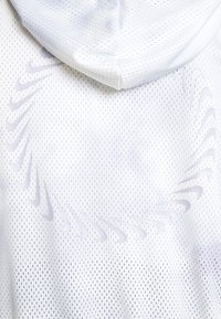 Bílá mikina s kapucí vyrobená z prodyšného síťovaného materiálu, s jemnými akcenty loga Nike a kapucí. Design zahrnuje texturovaný vzor ventilačních otvorů.