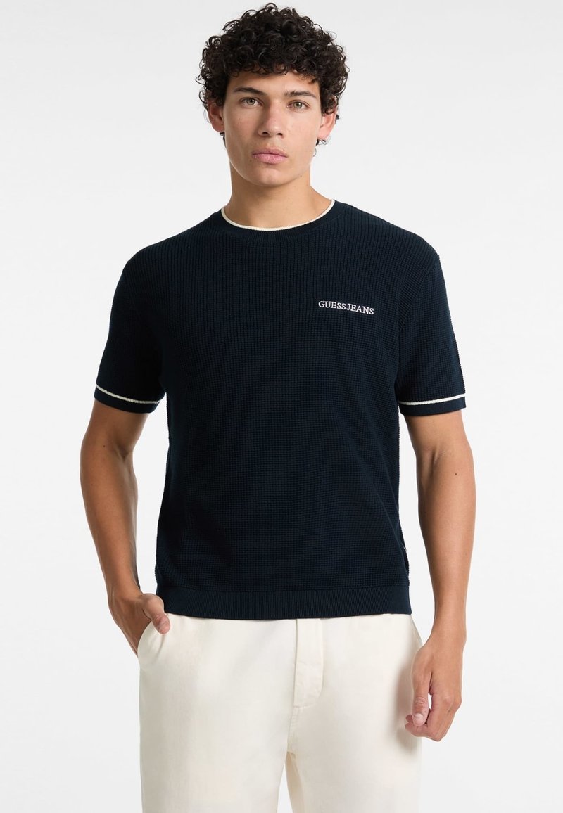 Tmavě modrá košile s krátkým rukávem s texturou, s bílými detaily a vyšitým logem 'GUESS JEANS' na levé hrudi. Nosit s světlými kalhotami.
