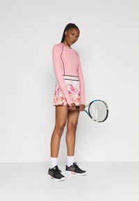 Mulher segurando uma raquete de ténis, vestindo uma blusa de manga longa rosa, uma saia de ténis floral, meias brancas e sapatilhas de desporto pretas, em pé contra um fundo liso.