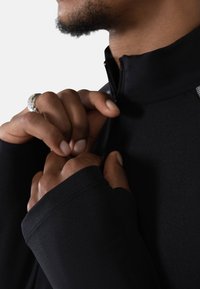 Svart, langermet topp med høy krage og glidelås. Stoffet ser glatt ut med tettsittende mansjetter. Hender som justerer glidelåsen.