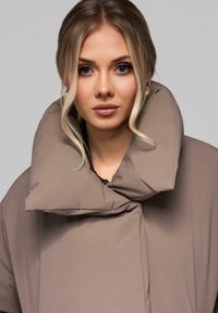 Kurtka puchowa w jasnym taupe, z dużym, pikowanym kołnierzem, gładką teksturą oraz z przodem zapinanym na sznurek.