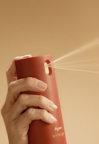 Dispositivo spray cilindrico rosso con finitura opaca, dotato di ugello dorato e un piccolo foro per la nebulizzazione, tenuto in mano contro uno sfondo beige.