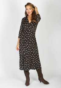 Kobieta z długimi włosami ubrana w czarną midi sukienkę w białe kropki i brązowe kozaki do kolan, pozująca z jedną ręką we włosach.