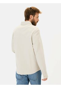 Lys beige skjorte med krave, lange ærmer og knapmanchetter. Glat tekstur med synlige sømdetaljer. Båret over blå jeans.