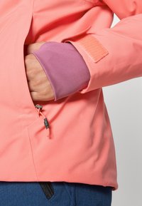 Korallos lágy héjú dzseki tépőzáras mandzsettával, lila bélésű ujjal, valamint zipzáras oldalzsebbel, narancssárga húzózsinórral a beállításhoz.