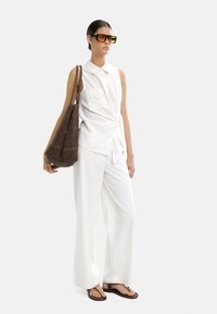 Biała bezrękawnikowa koszulka z wiązanym w pasie, z teksturowaną naszywką. Luźne białe spodnie i brązowa pleciona torba. Ciemne, oversize'owe okulary przeciwsłoneczne.