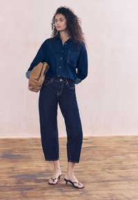 Jeansowa koszula w ciemnym odcieniu niebieskiego, z kieszenią i zapinana na guziki; zestawiona z krótkimi ciemnymi dżinsami i czarnymi klapkami na obcasie.