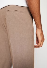 Brūnas faktūras bikses ar taisnu griezumu un sānu kabatas detaļu, attēlotas no aizmugures ar roku pie jostasvietas.