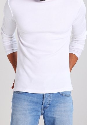 Homme portant une chemise blanche ajustée à manches longues et un jean bleu, les mains derrière le dos sur un fond uni.