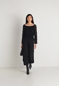 Μαύρη διάφανη μπλούζα με μακριά μανίκια και τετράγωνη λαιμόκοψη, συνδυασμένη με μαύρες παντελόνες με φαρδιά ποδιά. Το μοντέλο κρατάει μια τσάντα με σχέδιο μαύρες και άσπρες βούλες.