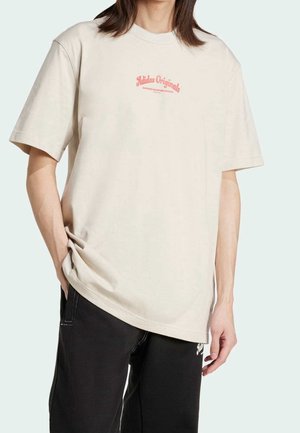 Osoba w beżowej koszulce Adidas Originals z czerwonym napisem i czarnych spodniach, stojąca z jedną ręką w kieszeni na jednolitym tle.