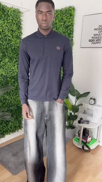 Ung mann i mørk langermet polo og brede gråfade jeans, som står i et rom med planter, kameraer og innrammet motiverende tekst.