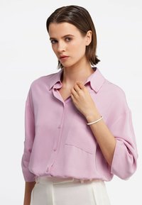 Camicia button-up rosa chiaro con una vestibilità rilassata, maniche corte e una singola tasca sul petto. Il modello indossa un bracciale semplice.