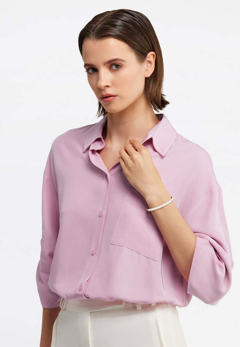 Camicia button-up rosa chiaro con una vestibilità rilassata, maniche corte e una singola tasca sul petto. Il modello indossa un bracciale semplice.