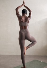 Woman in brown activewear performing tree yoga pose on green mat against beige wall indoors.