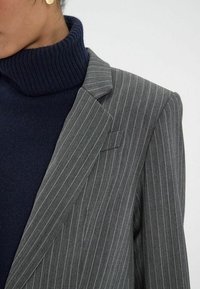Blazer gris à rayures avec revers en pointe, associé à un col roulé bleu marine. Le blazer présente une poche poitrine unique et une texture de tissu lisse.
