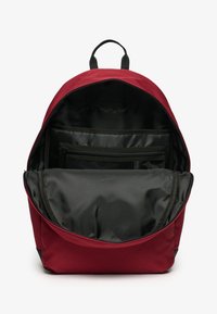 Mochila de tela roja con un gran interior negro. Cuenta con un bolsillo frontal con detalles de malla. Asa superior gruesa y cierre de cremallera.
