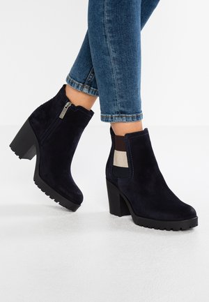 Person iført mørkeblå ruskind ankelsstøvler med blokhæle og lynlåse i siden, kombineret med opklemte blå jeans.